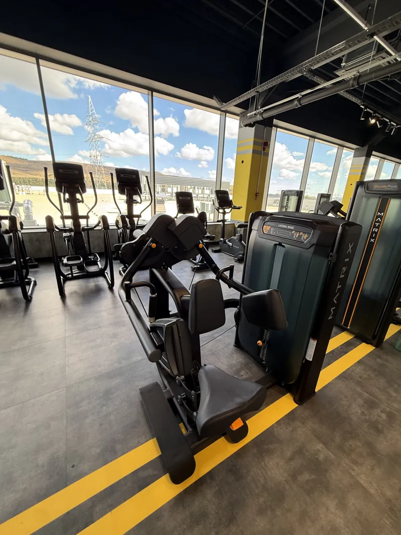 A chest press machine in front of tall gym windows